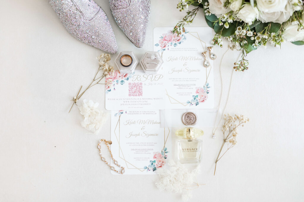 Flat lay details with invitation suite, flowers, perfume, shoes, and rings