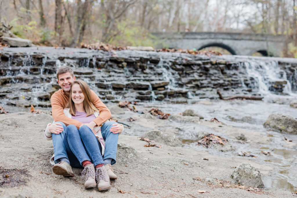 engaged couple sits on the rocky floor of the waterfall at sharon woods