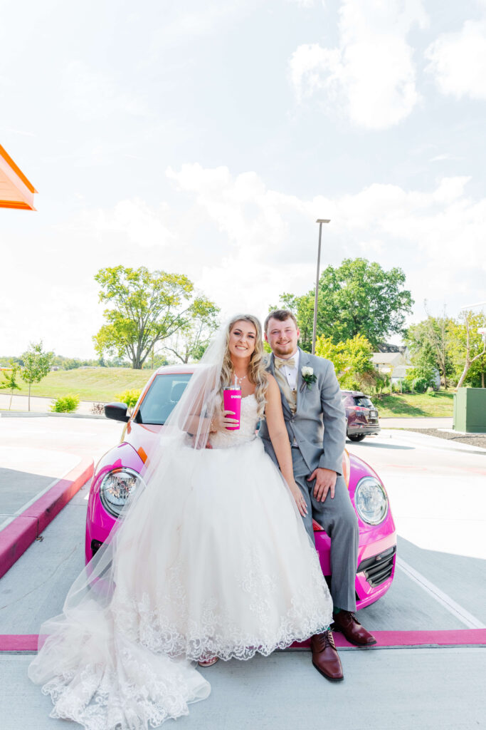 bride and groom at a dunkin donuts on their wedding day sitting on the a pink volkswagen holding a dunkin donuts cup