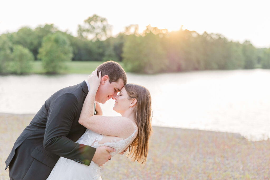 A bride and groom press their foreheads together and she has her hands wrapped around his head and he dips her back. The sun is setting behind them on Lake Lyndsay.
