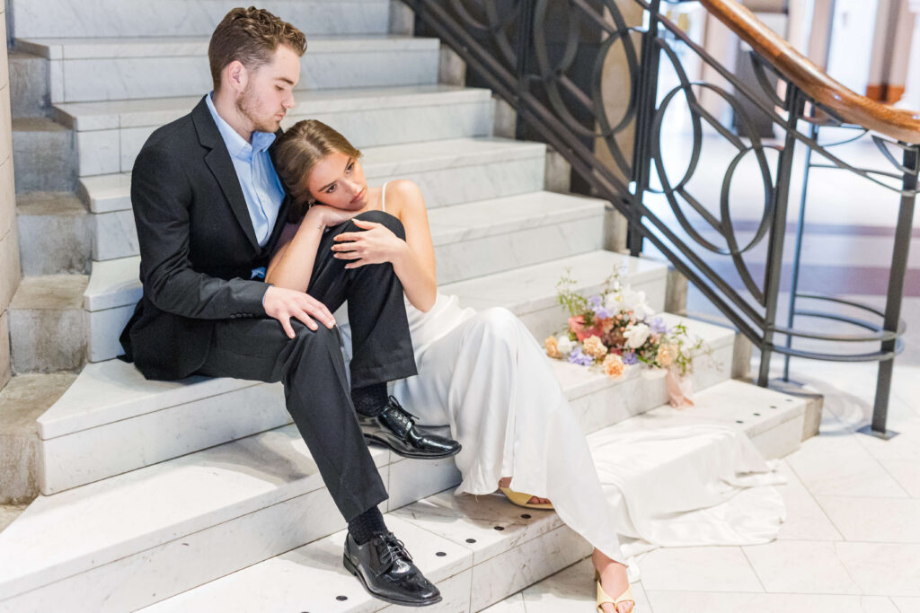 A bride and groom have their wedding at cincinnati art museum. They are sitting on the stairs as her head is in his lap and her bouquet is placed next to them. 