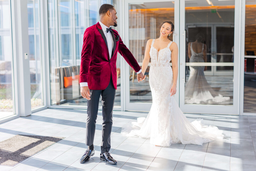 A groom in a maroon jacket and black pants, holds the hand of his bride as they walk through the summit hotel wedding. They are smiling, there are a lot of windows with sunlight coming in. 